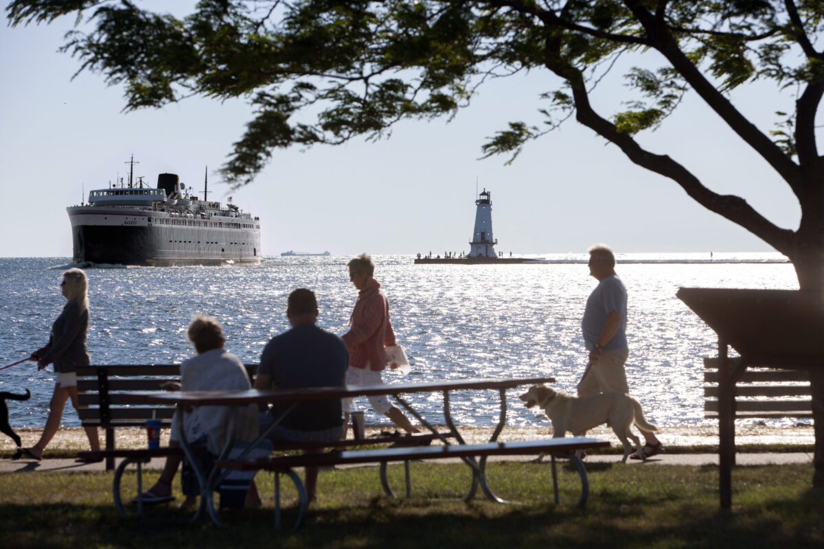 People stroll and picnic by the lakefront, with a boat and lighthouse in the background.
