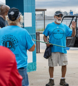 A volunteer wearing a shirt that says "Carlos Godinez Adopt-a-Beach Memorial Cleanup" speaks to volunteers near the shoreline.