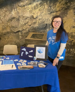 A smiling volunteer stands next to a table full of Alliance for the Great Lakes stickers, brochures, and other items.